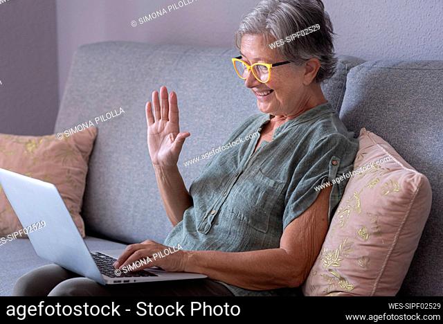 Smiling woman waving hand during video call through laptop at home