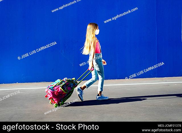 Blond girl walking with schoolbag on street against blue wall