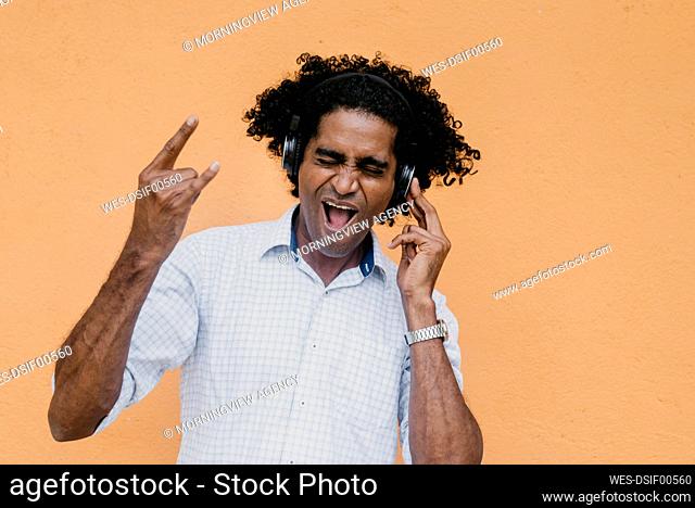 Man making horn sign while listening music on headphones in front of beige wall