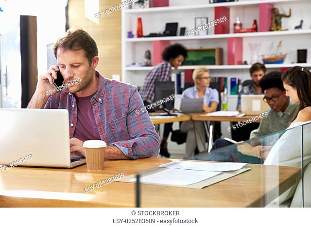 Businessman On Laptop Using Mobile Phone In Busy Office