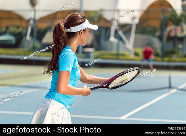 Side view of a beautiful and competitive woman smiling, while holding the tennis racket and the ball before starting the match on a professional tennis court