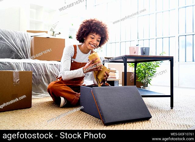 Smiling Afro woman keeping wrapped glasses in box while moving into new house