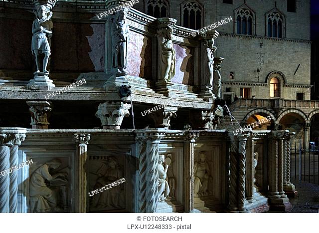 Detail, showing sculpted figures, of Fontana Maggiore Great Fountain in Piazza IV Novembre, Perugia with facade of Palazzo dei Priori Town Hall behind, at dusk