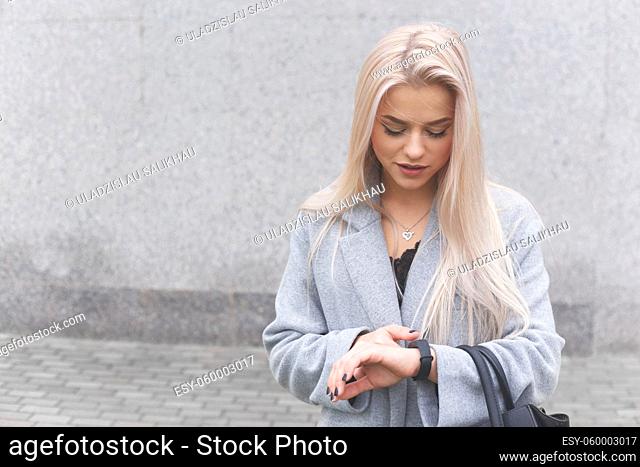 Portrait of young blondhair elegantly dressed woman in a coat uses a smart bracelet standing outdoors