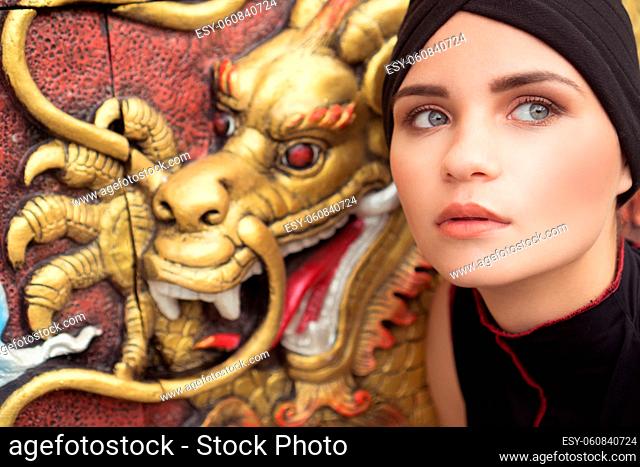 Fashion closeup portrait of young brunette woman with black hat over golden dragon on the wall background