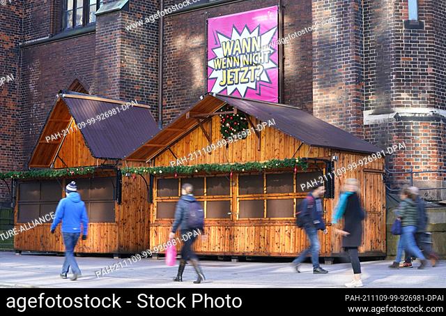 09 November 2021, Hamburg: Passers-by walk along wooden stalls from the planned Christmas market in the city centre. Above the wooden houses a poster with the...