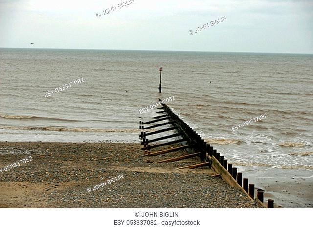 Wooden breakwater (groyne) sinking into the sea on a sandy beach with calm sea lapping against the shore. Blue sky with white cloud as background