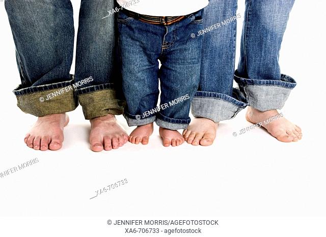 A family of three, mother, father and toddler son, stand on a white background wearing jeans. Only their legs and bare feet are shown