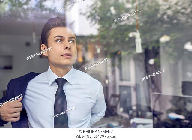 Portrait of young businessman behind glass pane in office putting on his jacket
