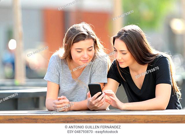 Two serious friends checking smart phone content sitting in a park
