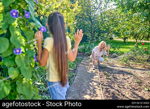 All excellent. Long-haired girl standing with her back to camera near climbing flowering plant greeting woman working in garden