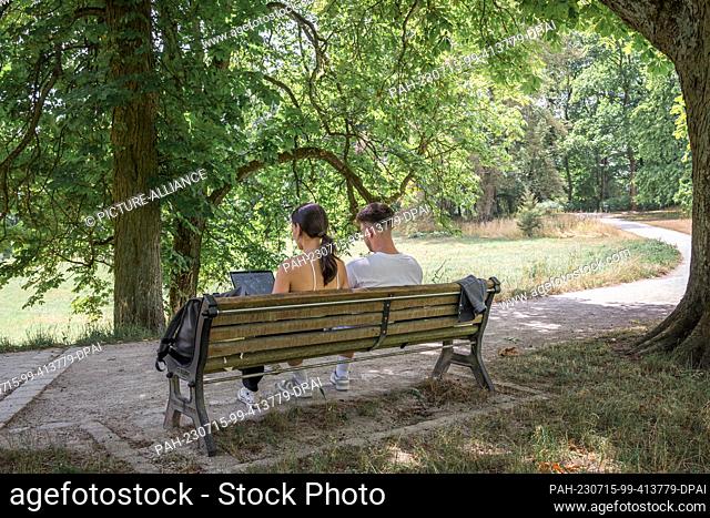 15 July 2023, Bavaria, Coburg: Two people are sitting in the shade on a bench in the courtyard garden. The person on the left is working on a laptop