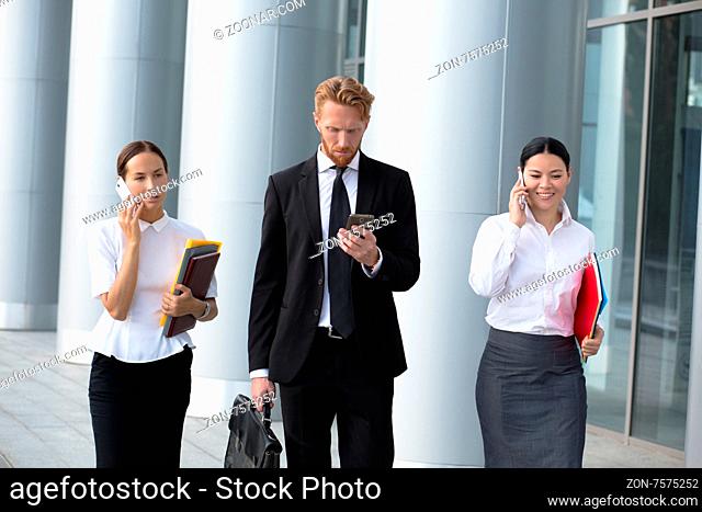 Businesswomen using cell phones while walking on path outside office. Man in black busines suit with brief case going to call to his secretary