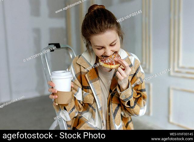 Young woman with disposable coffee cup eating food in apartment