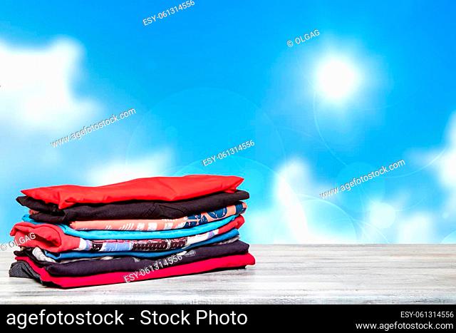 Stack colorful clothes. Closeup of a pile of colorful t-shirts or shirts on a bright table against abstract blurred sunny blue sky background