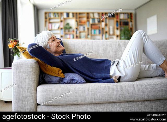 Smiling senior woman with hands behind head lying down on sofa at home
