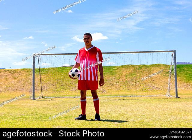 Portrait of Caucasian male football player wearing a team strip, training at a sports field in the sun, standing in front of a goal holding a football and...