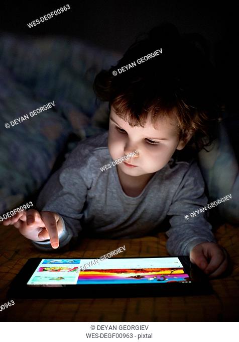 Little girl using digital tablet in bed under blanket
