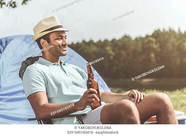 Delighted guy enjoying the nature while sitting on folding chair and holding bottle of beer. Forest and tent on background