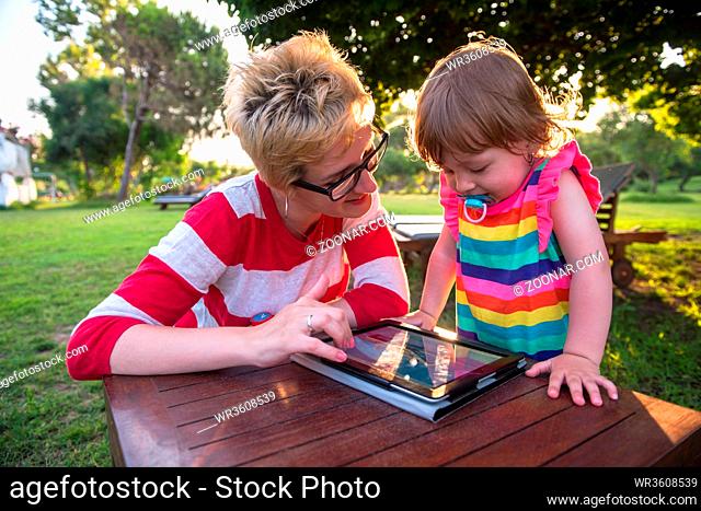 Happy mother and her little daughter enjoying free time using tablet computer while relaxing on holiday home garden during sunny day