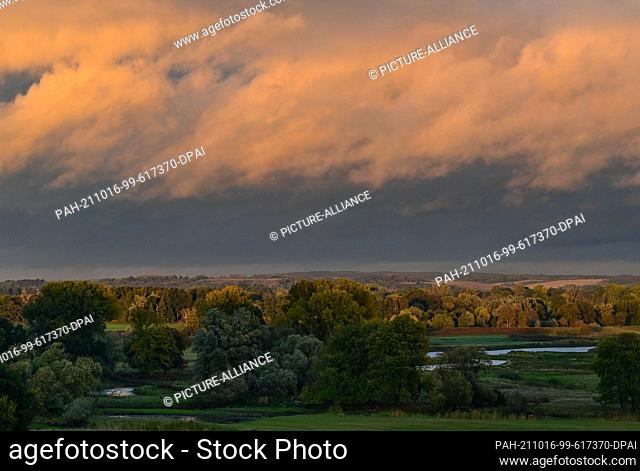 15 October 2021, Brandenburg, Lebus: Dark rain clouds are bathed in a warm light over the landscape on the German-Polish border river Oder by the light of the...