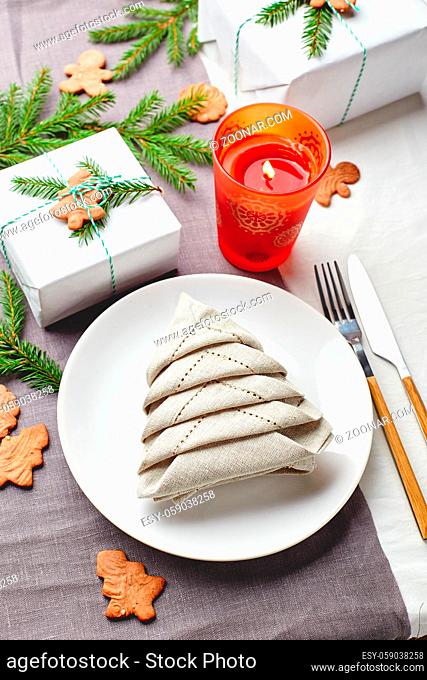 Christmas festive table setting, napkin in the form of Christmas tree on a plate on white tablecloth with gifts and decorations with fir sprigs and gingerbread...