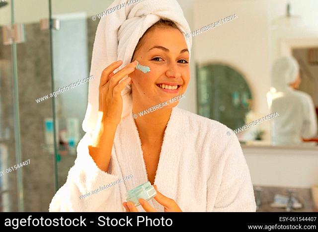 Portrait of happy caucasian woman wearing robe, applying beauty face mask in bathroom