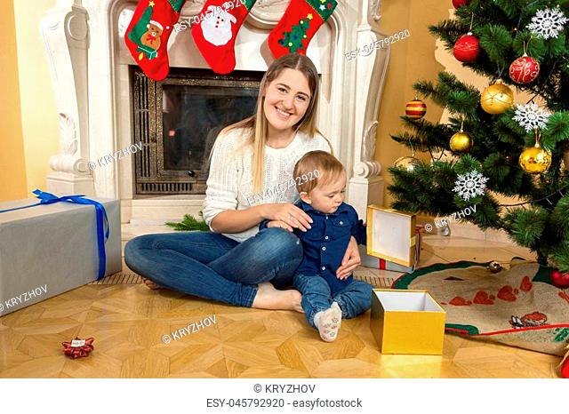 Baby boy with his mother opening gift boxes under Christmas tree at living room