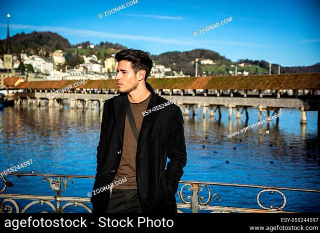Handsome man standing near metal fence in Lausanne. Famous wooden bridge and Water Tower on background