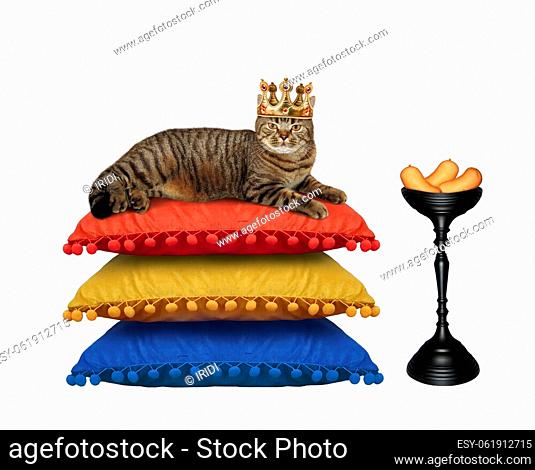 A beige cat in a crown is lying on a stack of pillows near a bowl with sausage. White background. Isolated