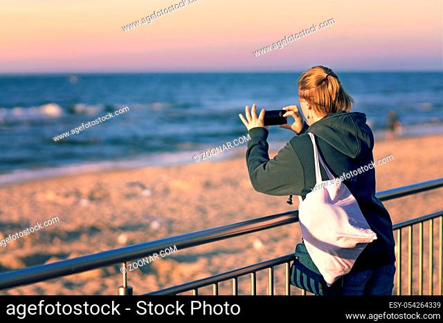 Blond Caucasian female taking pictures on her smartphone on a seafront promenade in a polish resort town Sarbinowo, Poland