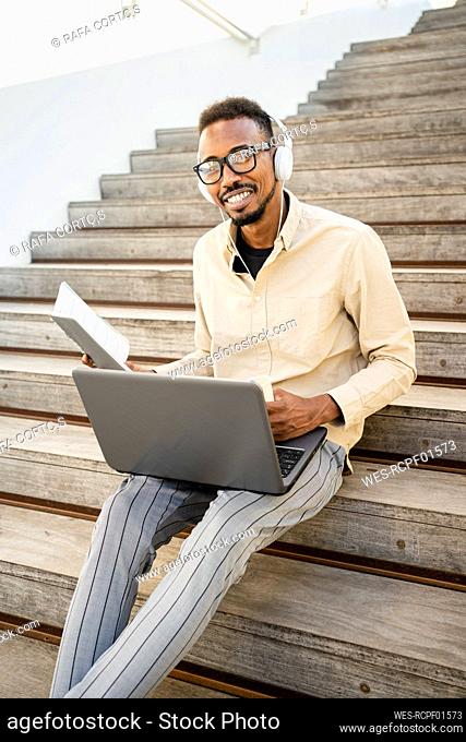 Happy man sitting with laptop wearing headphones on steps