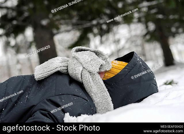 Boy covering face with gray scarf lying on snow in winter