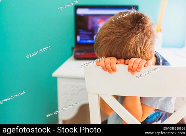 The sad little boy sits in front of laptop and doesn't want to look lesson. Concept of boring learning at home