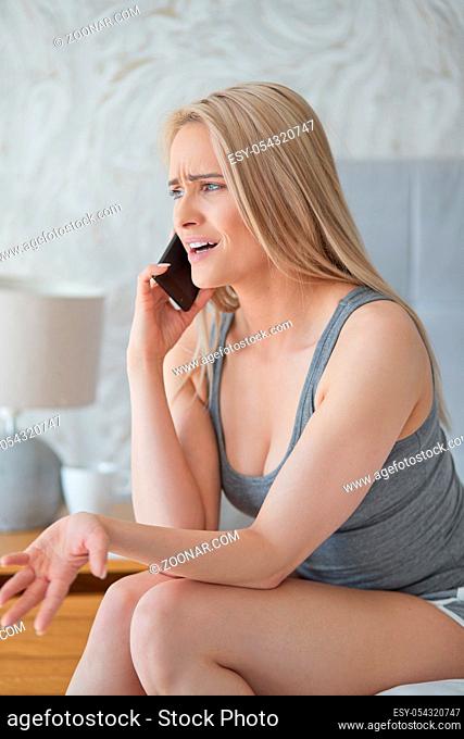 Middle aged blond woman sitting in her home bed and arguing with someone on mobile phone
