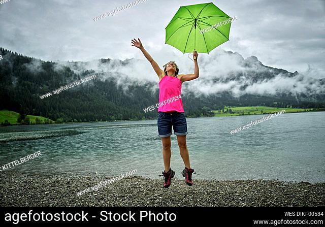 Happy mature woman holding umbrella while jumping at Haldensee lakeshore during rainy season