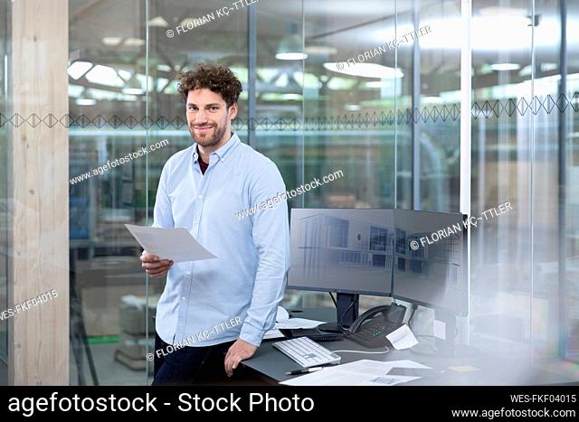 Smiling male entrepreneur with business strategy standing by desk against glass wall in factory