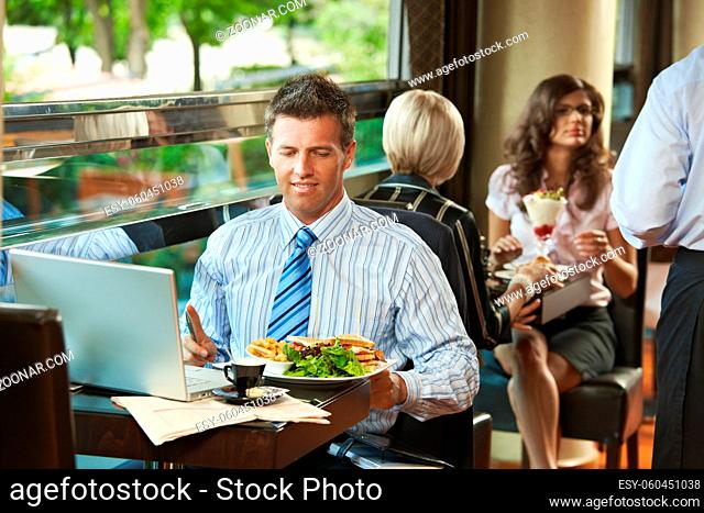 Businessman sitting at table in cafe, eating club sandwich and using laptop computer. Waiter serving sweets in the background