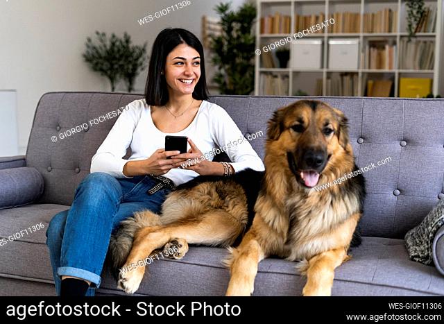Happy woman with smart phone looking away while sitting with dog on sofa at home
