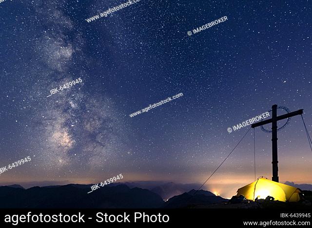 Night sky with stars and milky way over Lechtal mountains, in the foreground summit cross of the Geierkopf with tent, Reutte, Ammergau Alps, Tyrol, Austria