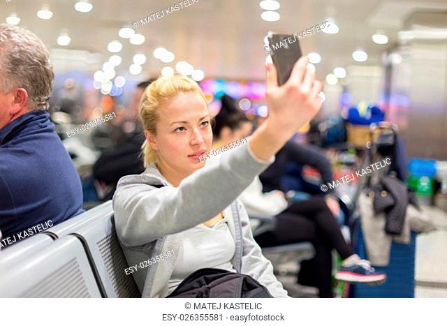 Casual blond young woman taking selfie photo using her cell phone while waiting to board a plane at the departure gates