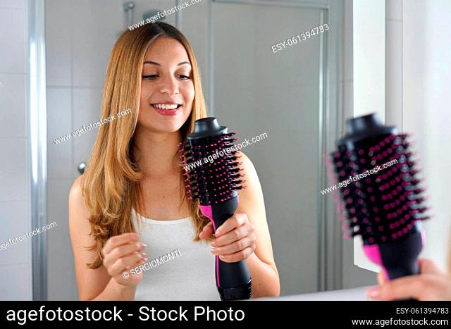 Smiling girl holds round brush hair dryer in her bathroom at home. Young woman looking satisfied her salon one-step brush hair dryer and volumizer