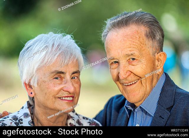 Head-and-shoulder portrait of an 80 year old retired couple in front of a blurred green background looking friendly into the camera