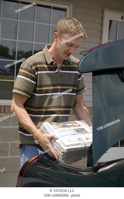 Newspaper delivery person loading newspapers into trunk of car
