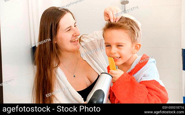 Young mother blowing with air from hairdryer on her little son singing in haircomb after having bath. Concept of child hygiene and health care at home