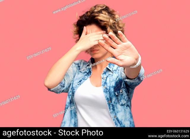 Stop, I don't want to look at this. Portrait of confused or scared young woman with curly hair standing covering her eyes and showing stop sign