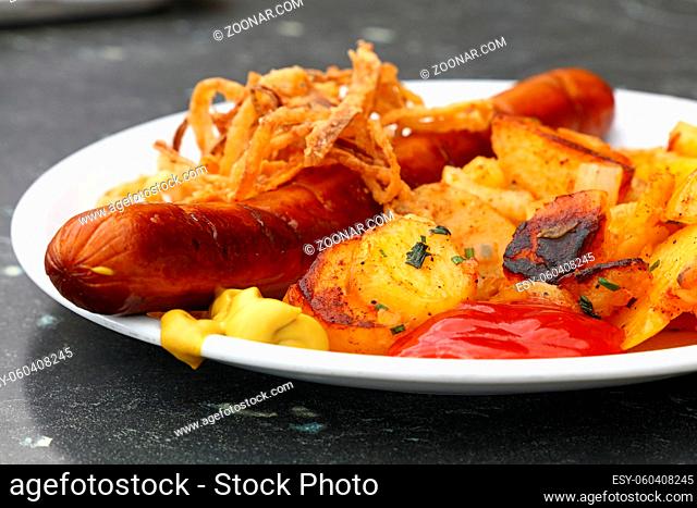 Close up portion of one big grilled sausage with homemade roasted potato, fried onion rings, ketchup and mustard on white plate over grey table, low angle view