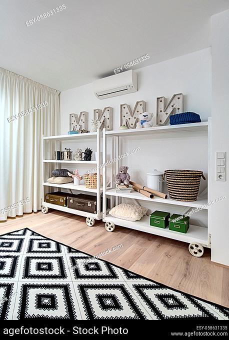 Children room in a modern style with white walls and a parquet with a carpet on the floor. There is white shelves on the wheels with toys, boxes, pillows
