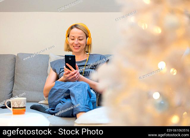 Young cheerful woman sitting indoors at home living room sofa using social media on phone for video chatting and staying connected with her loved ones