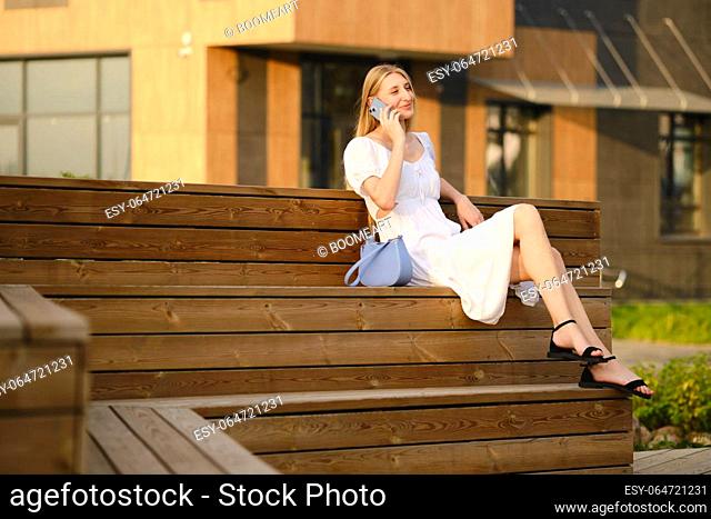 Pleased young woman sitting on bench and talking on smartphone in warm evening